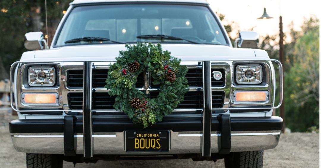 A White Dodge Pickup Truck with a Green Wreath on the Grill and California License Plate that reads Bouqs Ready for Holiday Travel