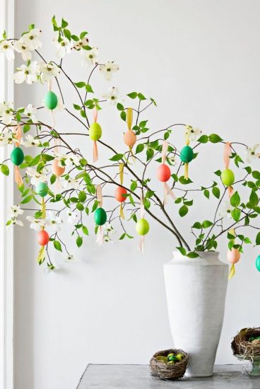 Small tree branches protruding from white vase with Easter eggs hanging from the branches