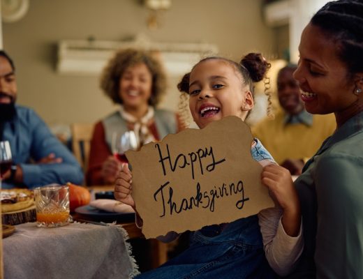 Little girl holding a sign that reads "Happy Thanksgiving" at a table with her family.