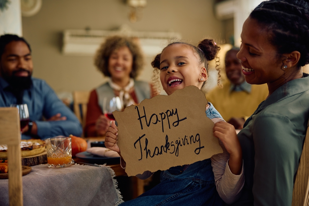 Little girl holding a sign that reads "Happy Thanksgiving" at a table with her family.