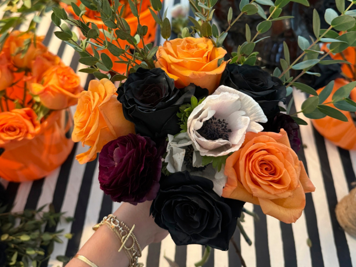 A hand holding out an arrangement of white, orange, and mauve flowers.