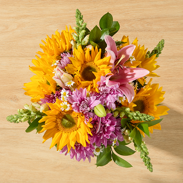 Yellow sunflowers with a pink lily, lavender cushion poms, white feverfew, and purple snapdragons.