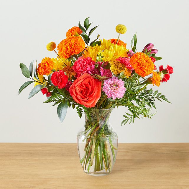 A clear vase flower arrangement with orange roses, marigolds, and carnations.