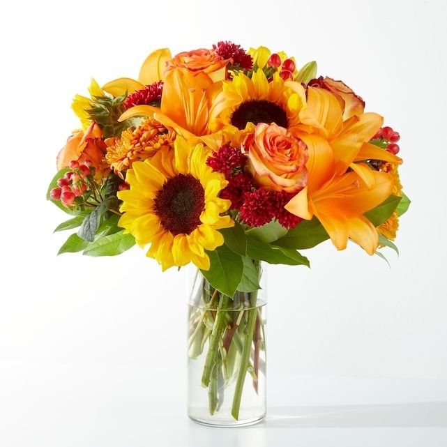 An arrangement with yellow sunflowers, orange lilies and orange roses in a clear vase.